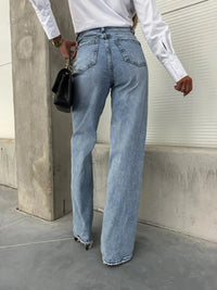 Person wearing light blue jeans and a white shirt against a concrete wall.
