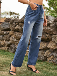 Person wearing blue jeans with slight distressing, standing against a stone wall.