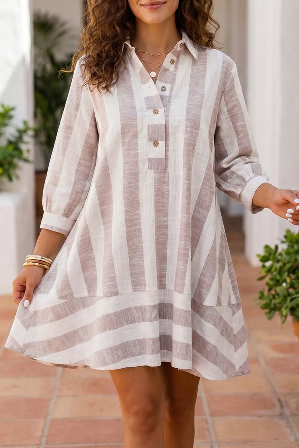 A woman wearing a loose, long-sleeve striped shirt dress in beige and white, standing in a sunlit outdoor corridor with potted plants.