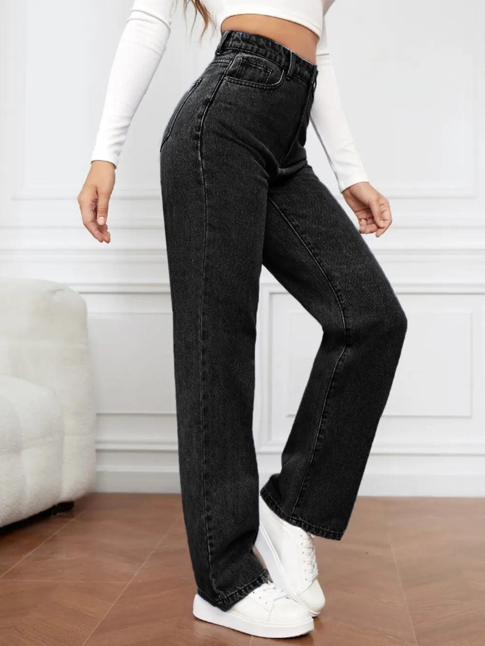 Person wearing black high-waisted jeans and white sneakers in a minimal indoor setting.