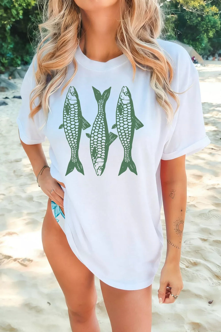 Woman wearing a white t-shirt with green fish graphics on a beach.