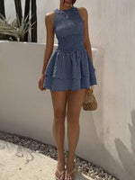 Woman wearing a blue checkered dress standing against a white wall with plants in the background.
