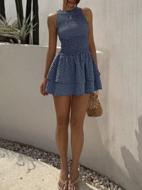 Woman wearing a blue checkered dress standing against a white wall with plants in the background.