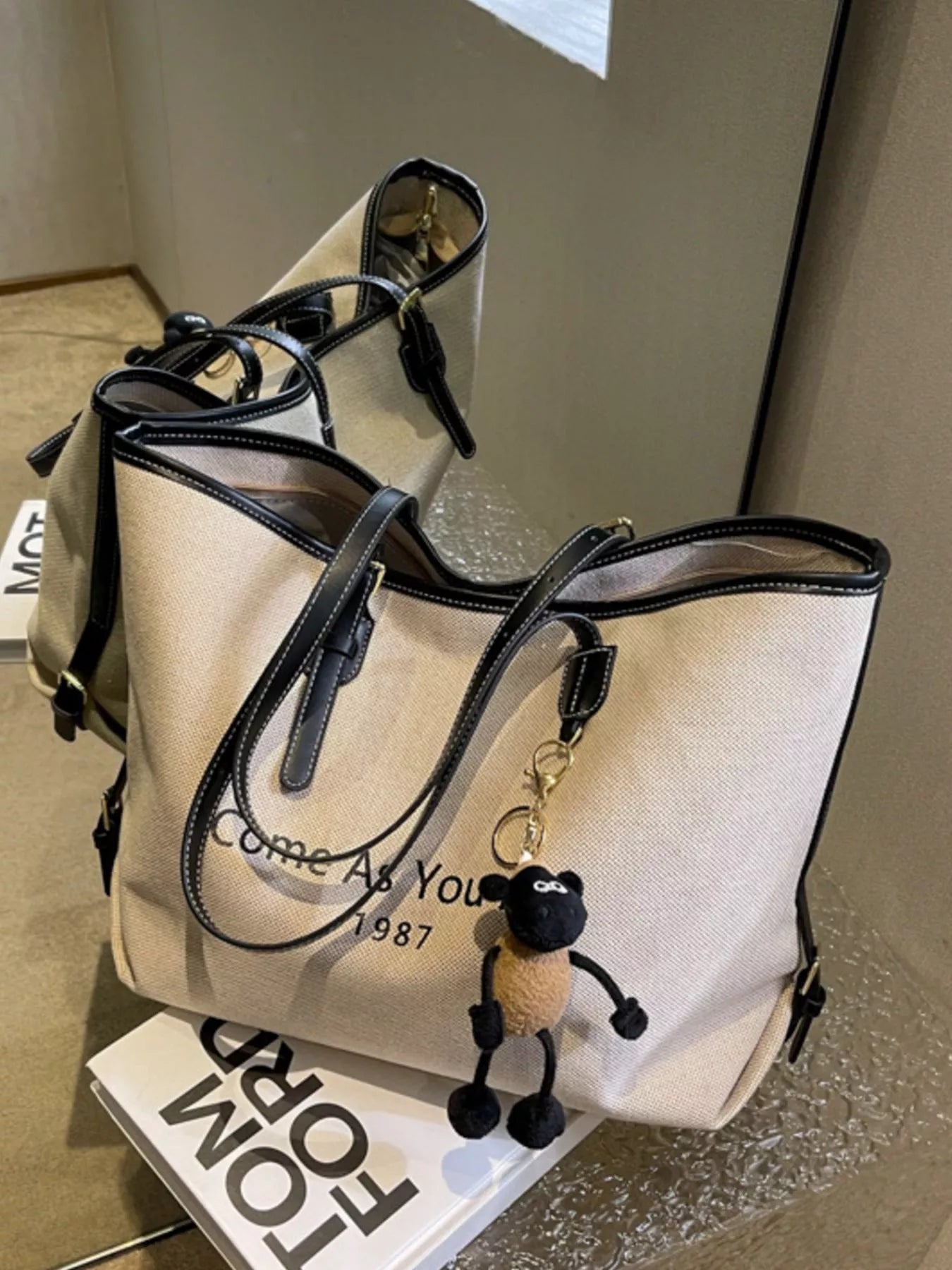 Beige handbag with black accents and a keychain on a reflective surface.