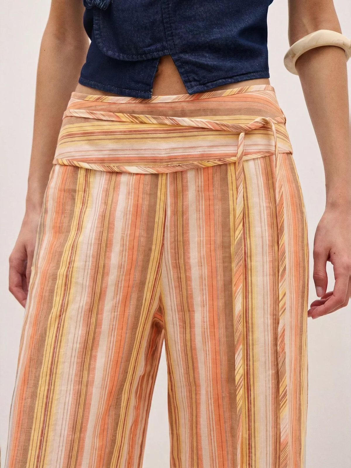 A close-up of a model wearing high-waisted, wide-leg trousers with vertical stripes in orange, yellow, and beige, paired with a dark denim top.