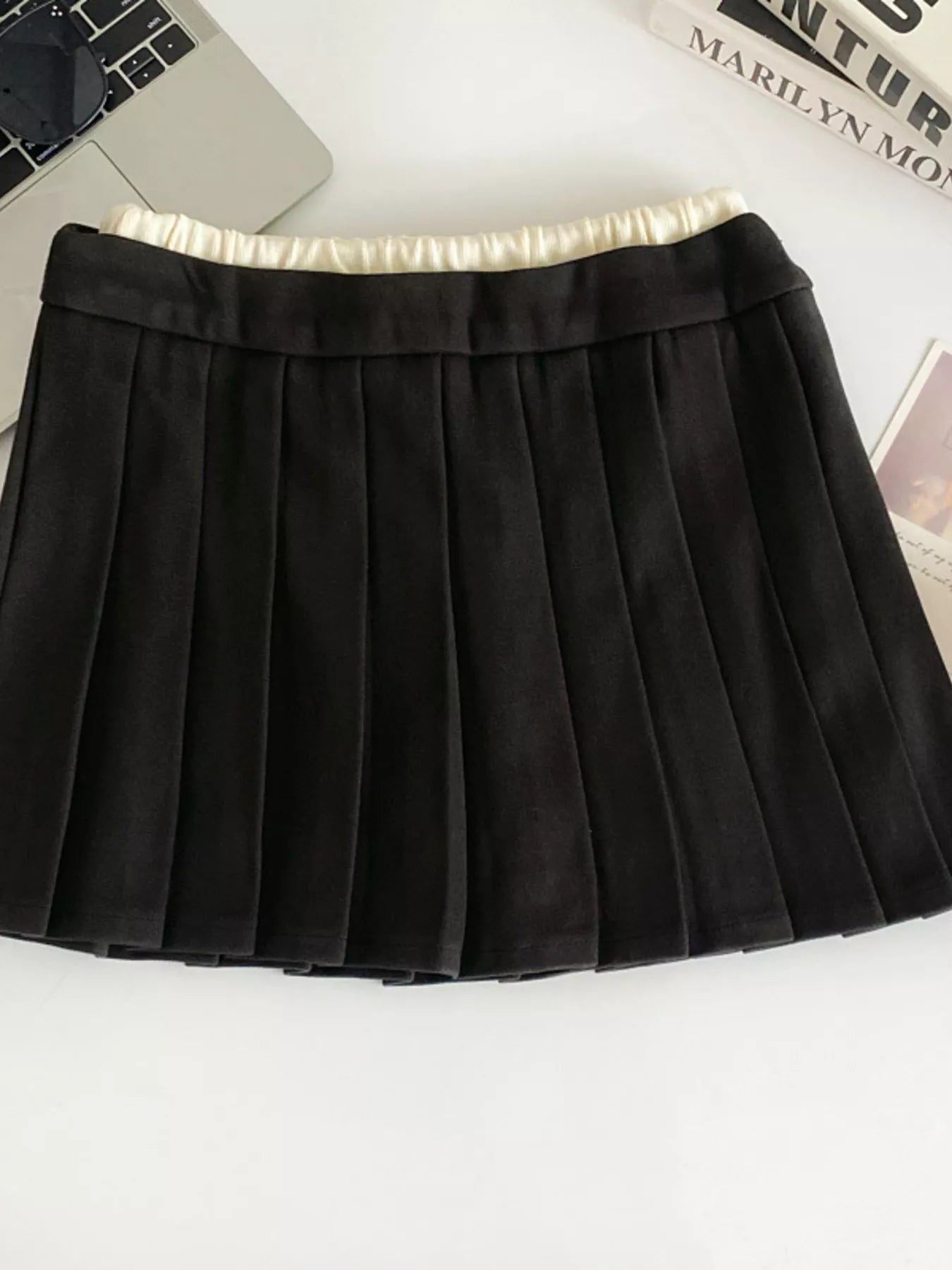Black pleated skirt on a white surface with a laptop and magazine in the background.