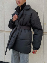 Person wearing a black puffer jacket with a belt against a light-colored wall.
