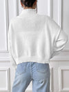 Person wearing a white sweater and light blue jeans against a white paneled wall.