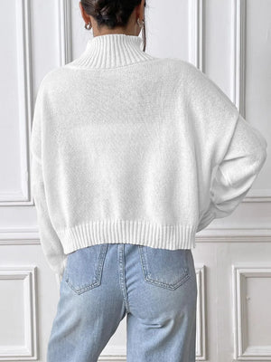 Person wearing a white sweater and light blue jeans against a white paneled wall.