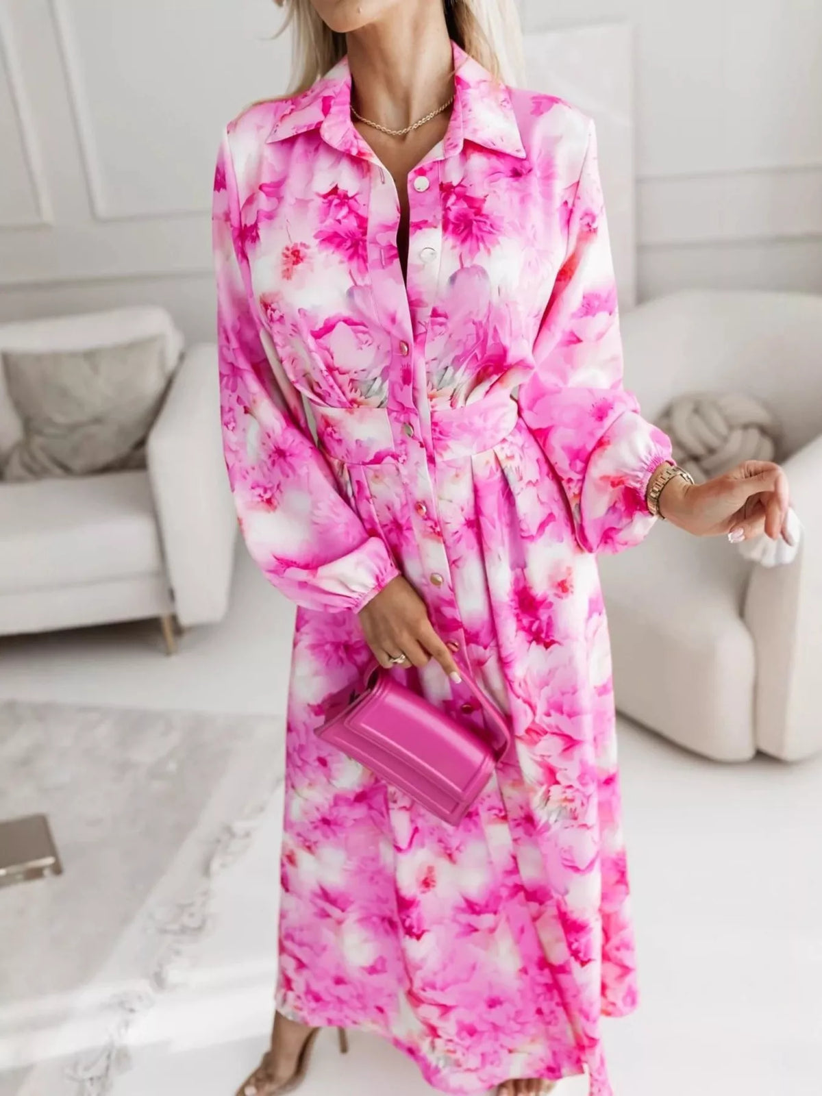 A woman wearing a long-sleeve pink floral shirt dress with a tie waist, holding a matching pink clutch bag in a bright, modern living room setting.