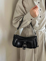 A person wearing a light beige long-sleeve button-up dress carries a black faux-croc leather shoulder bag with silver hardware against a plain beige wall.