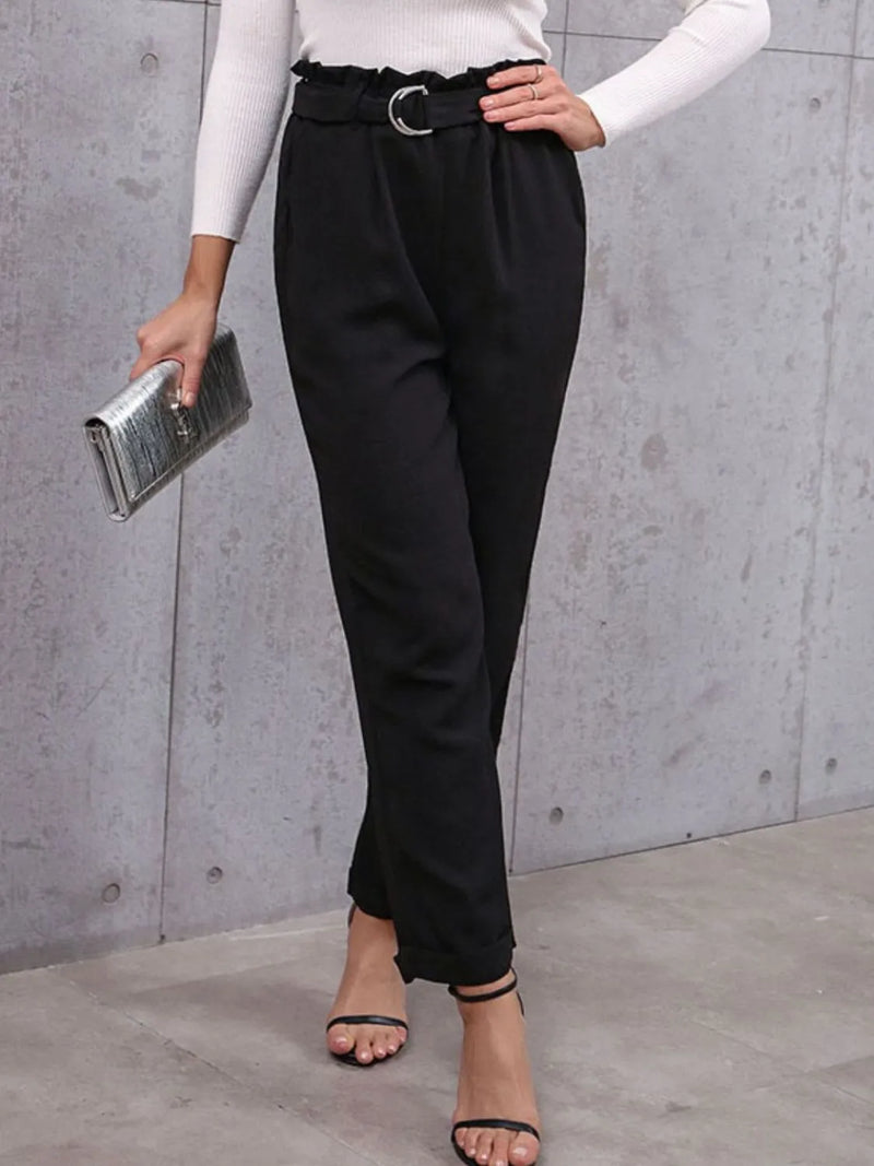 Person wearing black pants and a white top, holding a clutch against a gray wall.