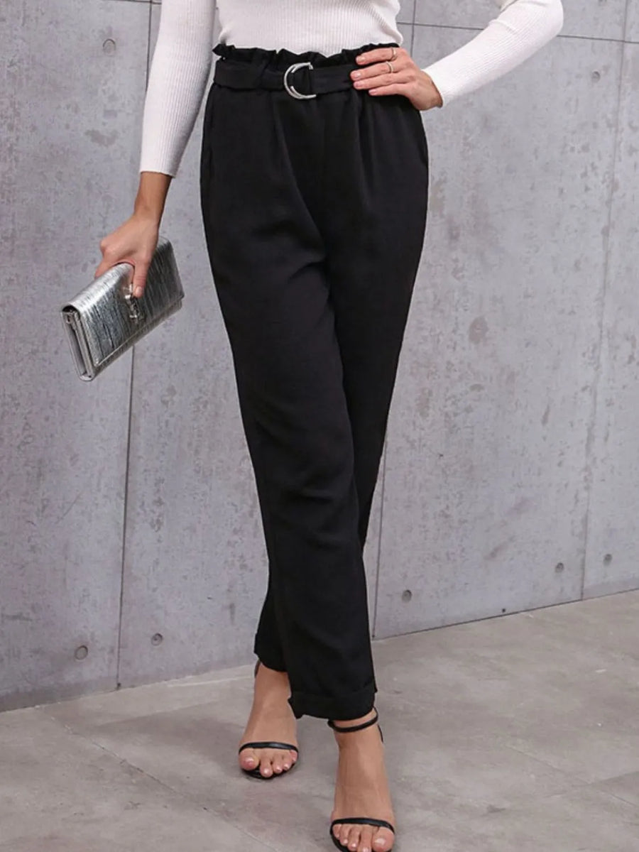 Person wearing black pants and a white top, holding a clutch against a gray wall.