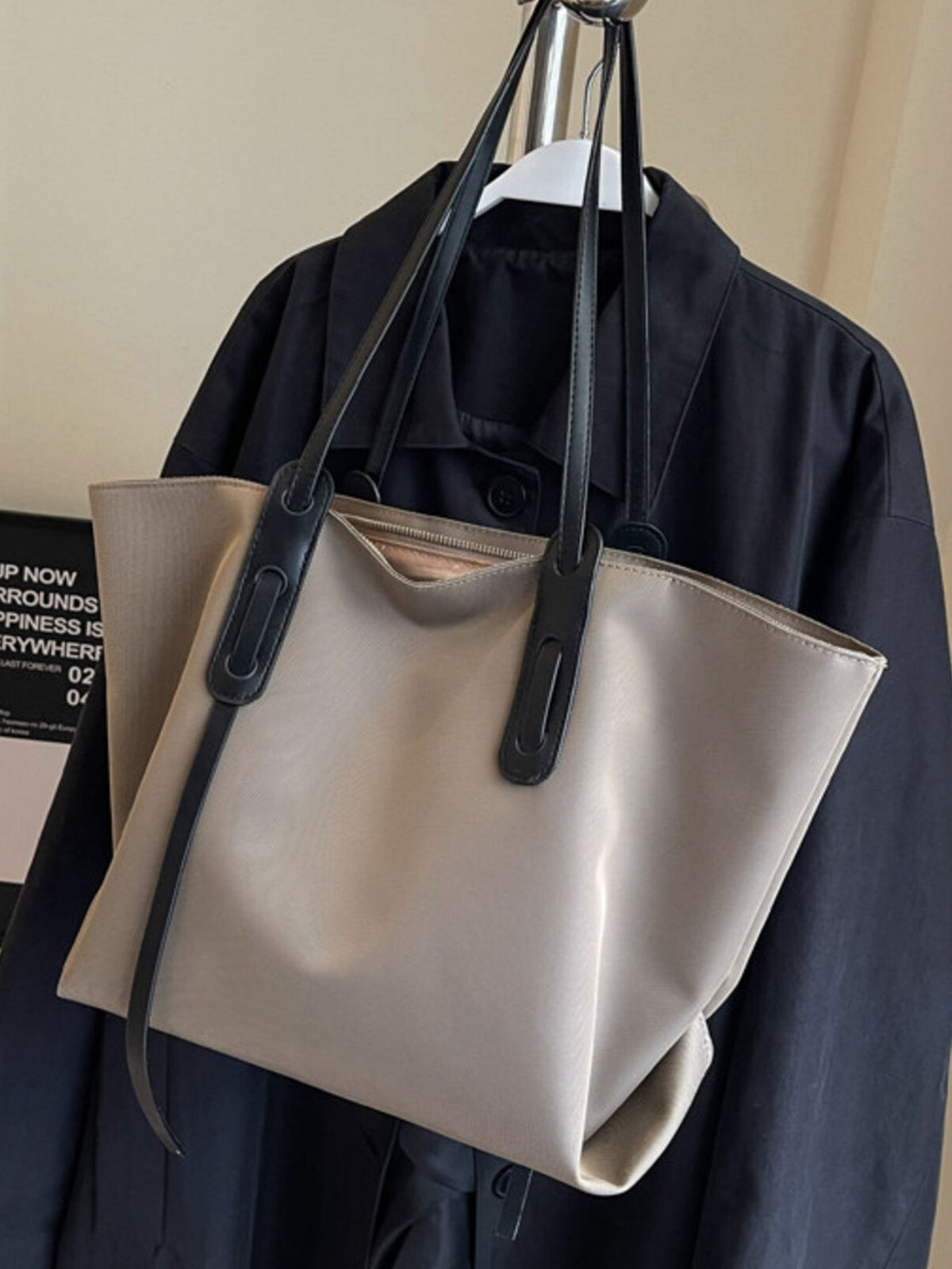 Beige tote bag with black straps hanging on a dark coat against a neutral background
