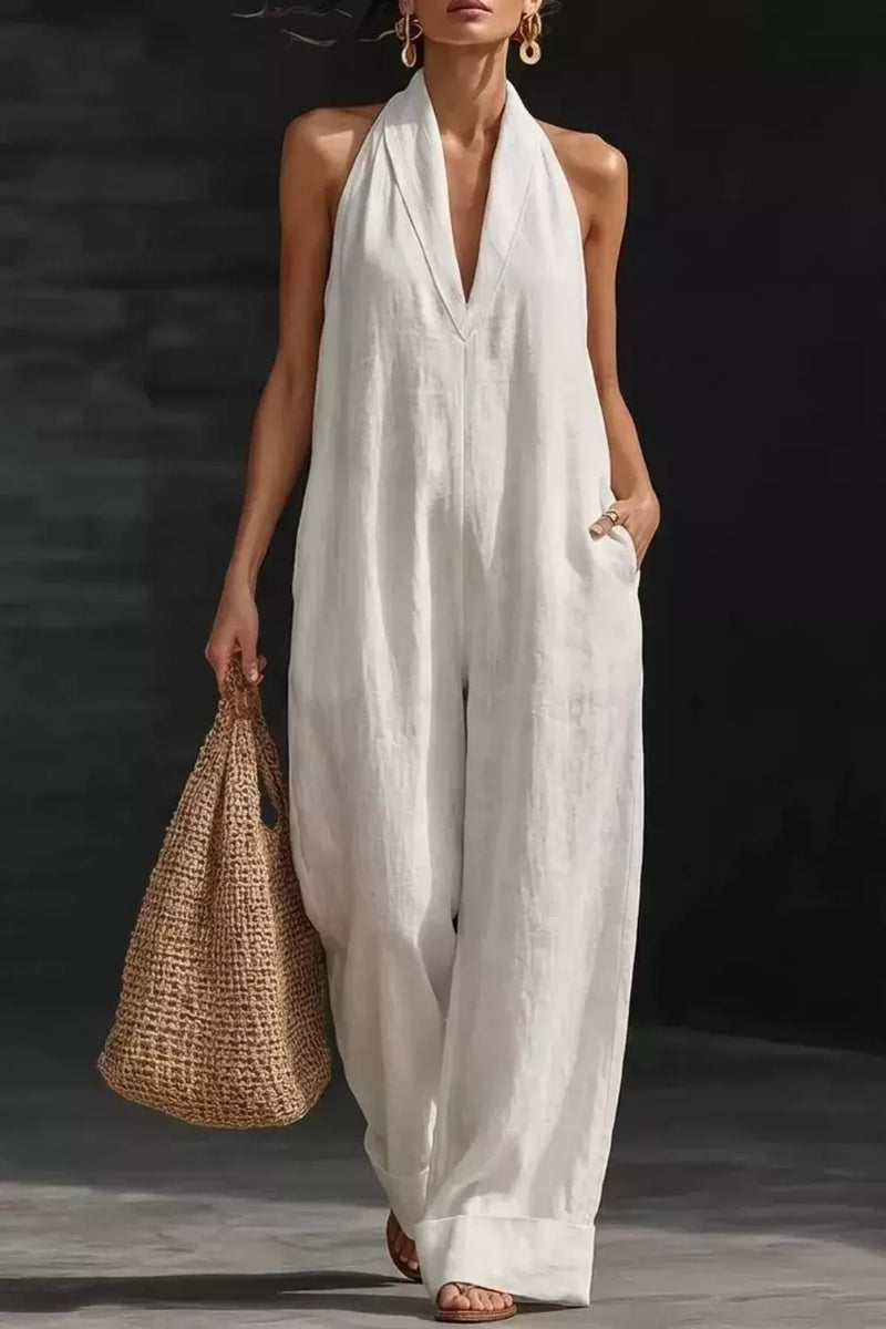Woman wearing a white halter neck jumpsuit holding a woven bag against a dark background