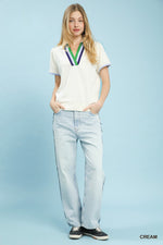 Woman wearing a white polo shirt with colorful accents and light blue jeans on a light blue background.