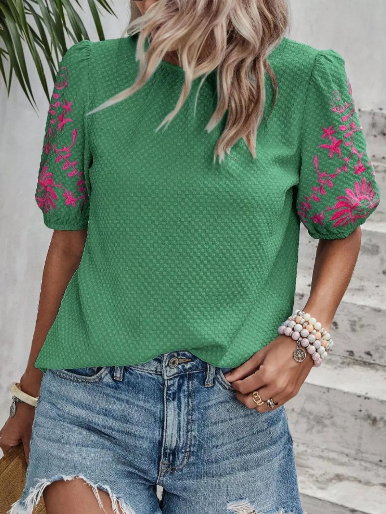 Person wearing a green blouse with pink floral embroidery and denim shorts.
