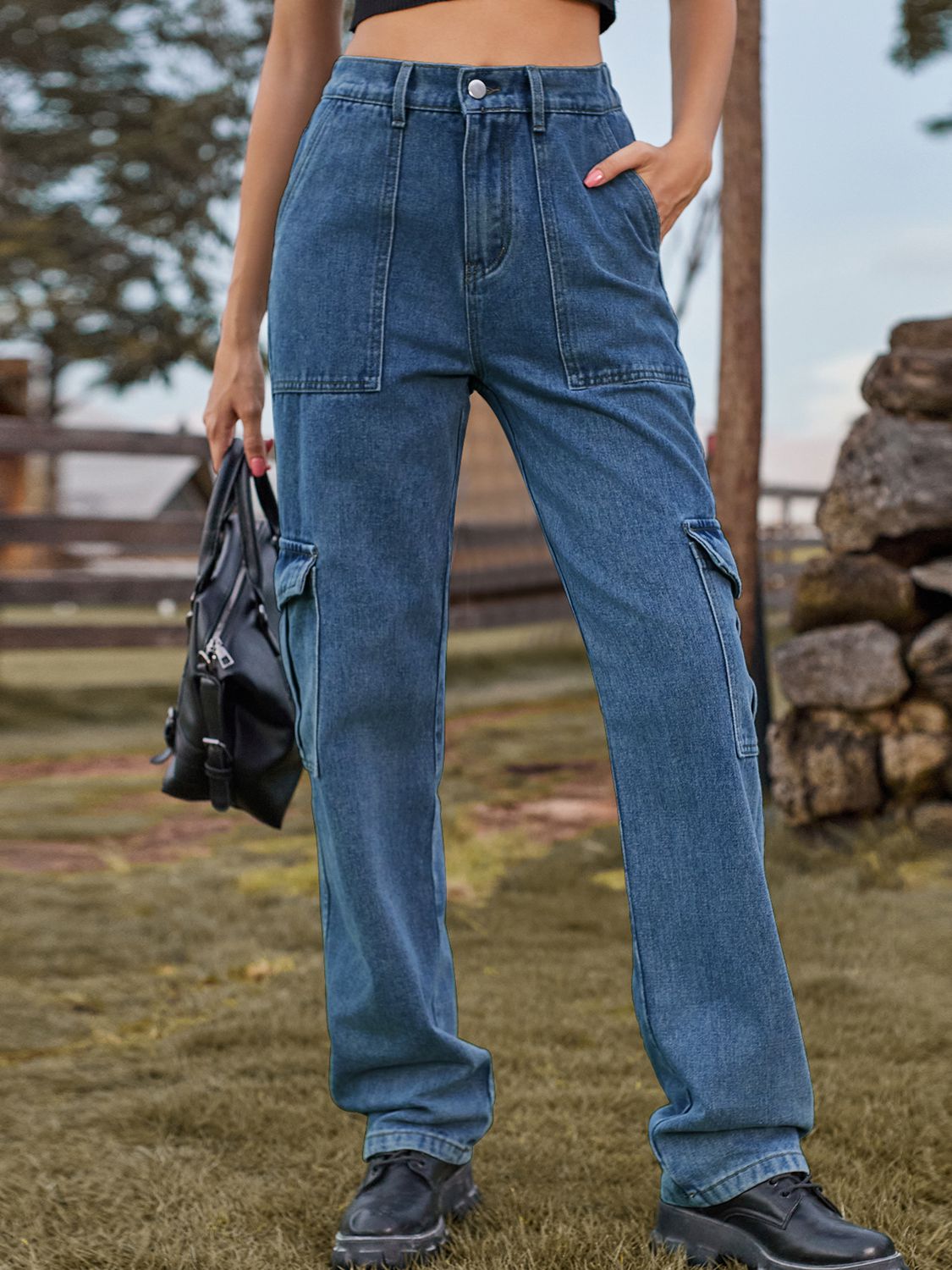 Person wearing blue jeans holding a black bag outdoors with a stone wall and trees in the background.