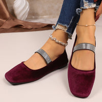 Burgundy velvet flats with crystal straps worn by a person on a neutral background