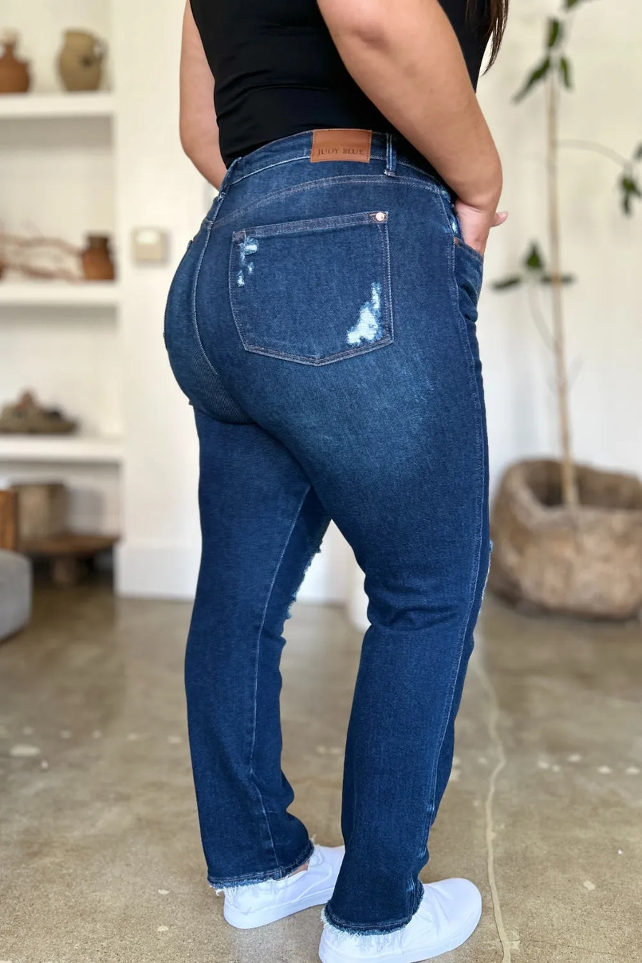 Person wearing blue jeans and white sneakers in a room with a shelf and plant in the background