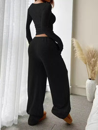 Person wearing a black ribbed outfit in a room with white curtains and a vase with dried plants.