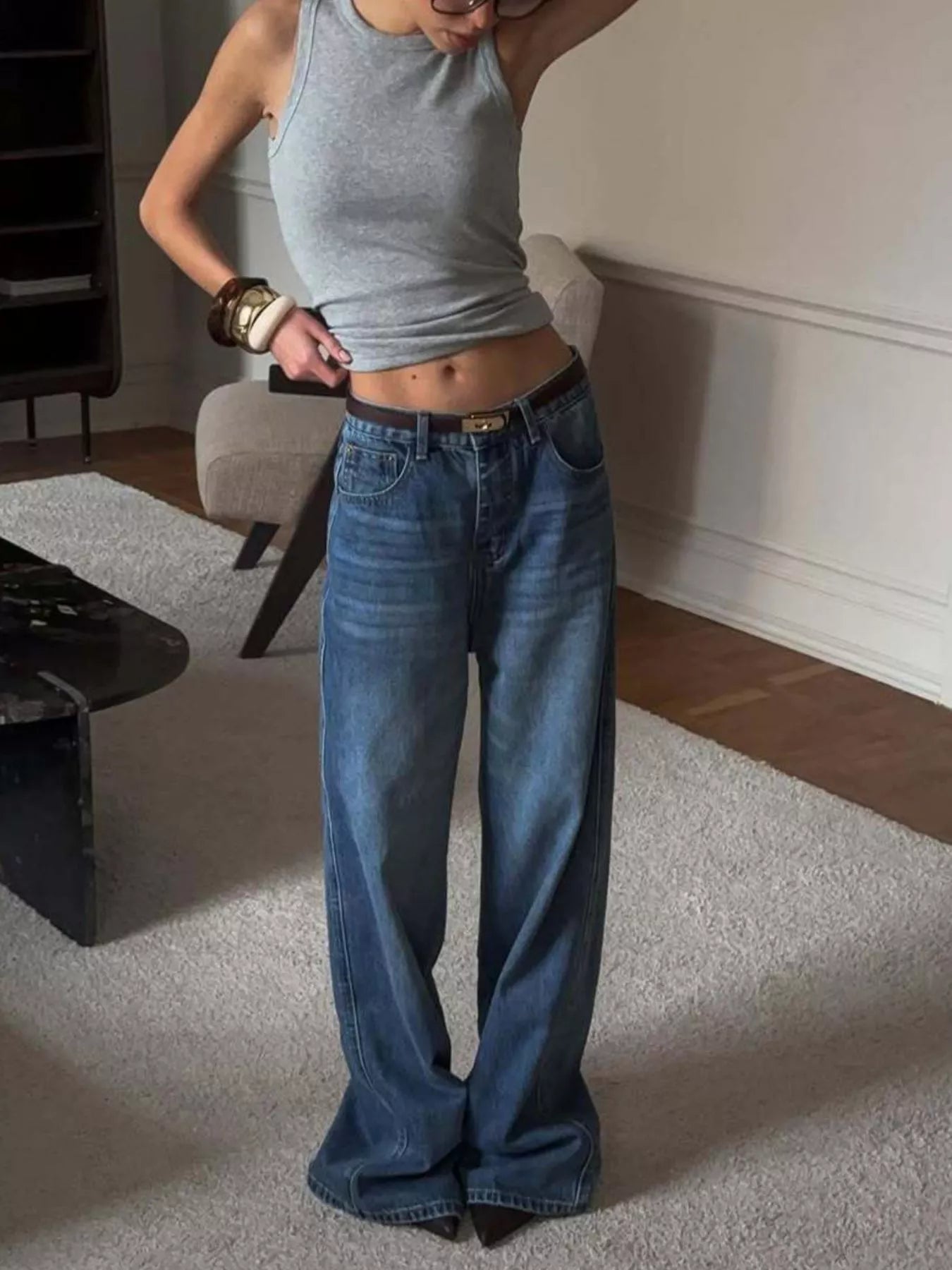 Person wearing a gray tank top and blue jeans in a room with a rug and furniture.