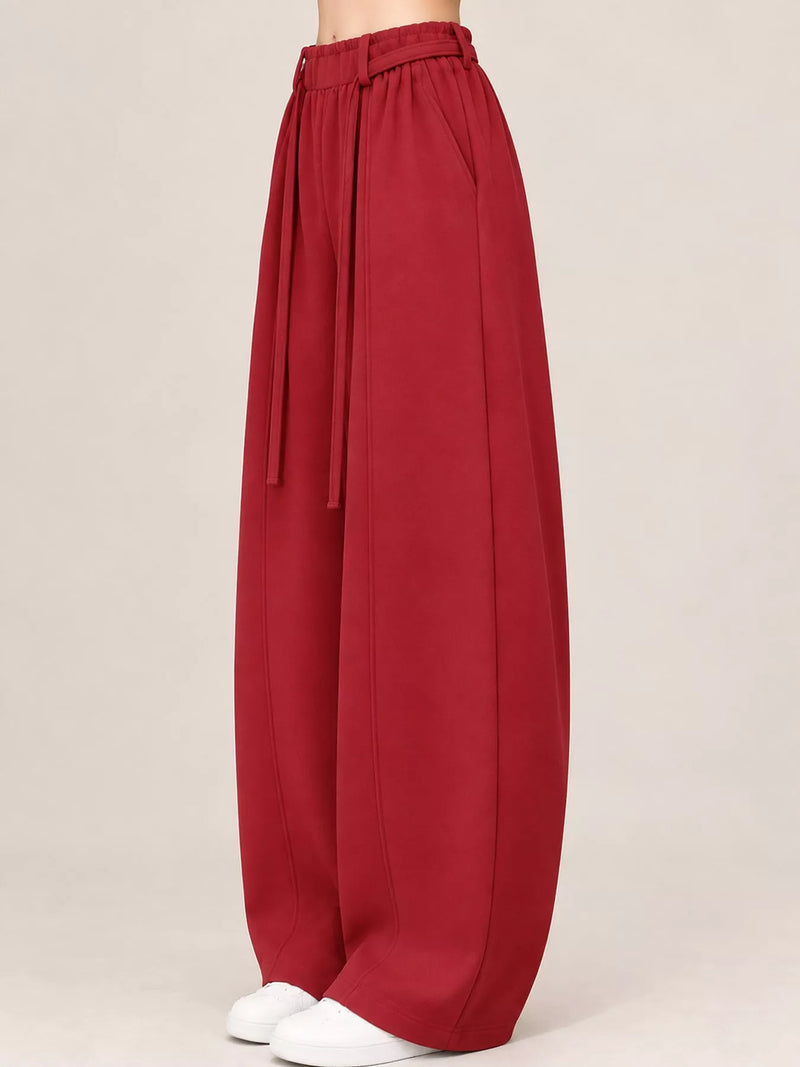 A pair of wide-leg, balloon-style pants in a deep red or burgundy color, featuring an elastic waistband with a matching tie belt, shown on a model from the waist down against a light beige studio background.
