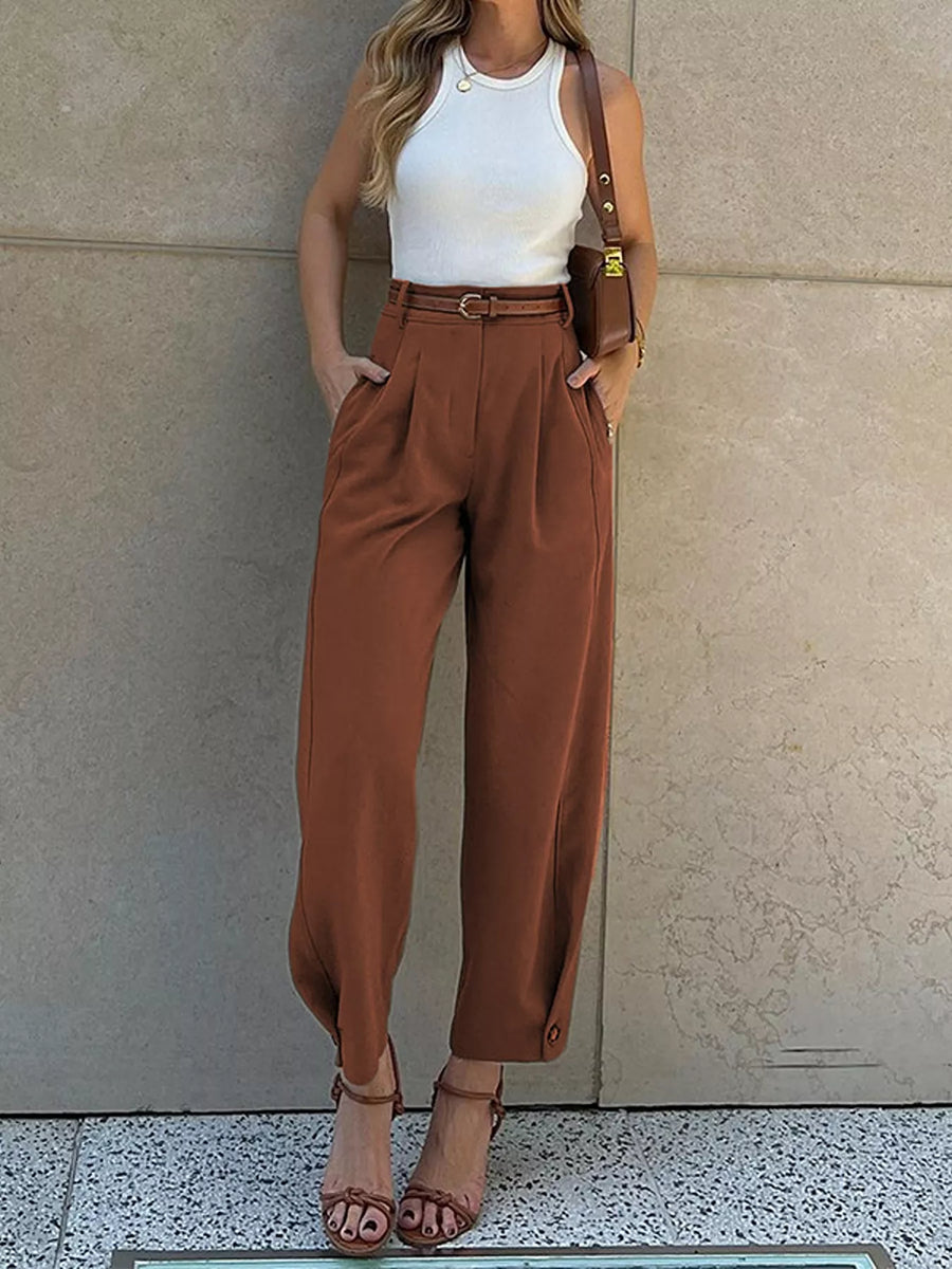 A woman models a pair of high-waisted, wide-leg brown trousers paired with a white ribbed tank top, brown leather sandals, and a brown shoulder bag, standing against a beige stone wall.