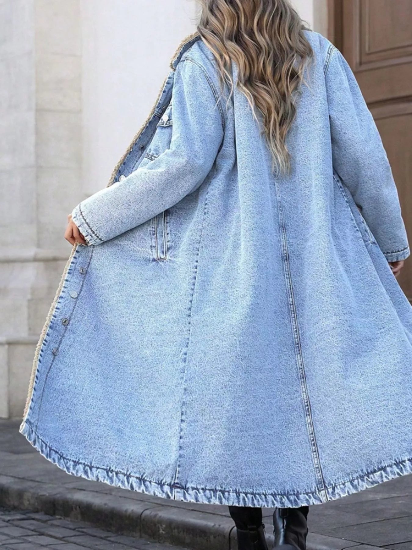 Person wearing a long denim coat standing on a sidewalk.