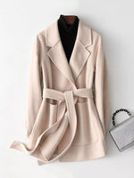 A light beige belted coat with wide lapels, styled on a hanger over a black turtleneck and accompanied by a small silver textured handbag on a white background.