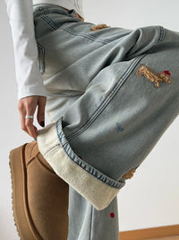 Light blue jeans with embroidered designs worn by a person, paired with brown shoes on a light gray background.