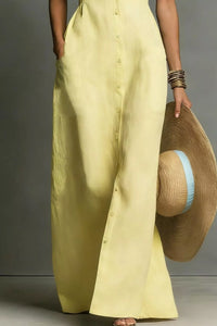 Person wearing a light yellow button-up shirt and wide-leg pants, holding a straw hat against a neutral background.