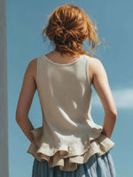 Woman wearing a beige sleeveless top with ruffled layers against a blue sky.