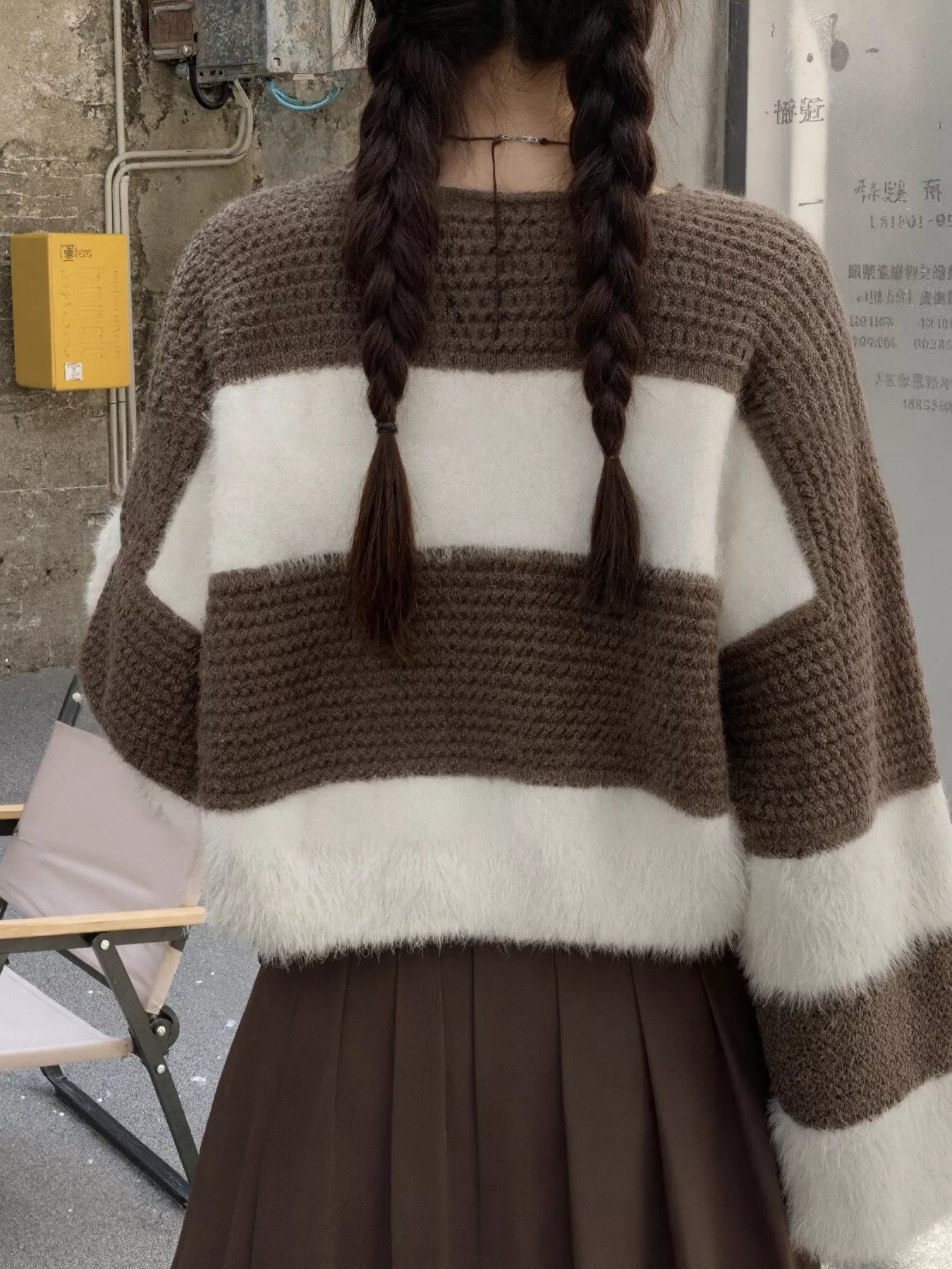 Person wearing a brown and white striped sweater with braided hair, standing against an urban background.