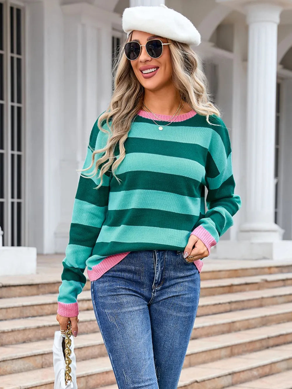 Woman wearing a green and pink striped sweater with jeans and a white hat, standing on steps.