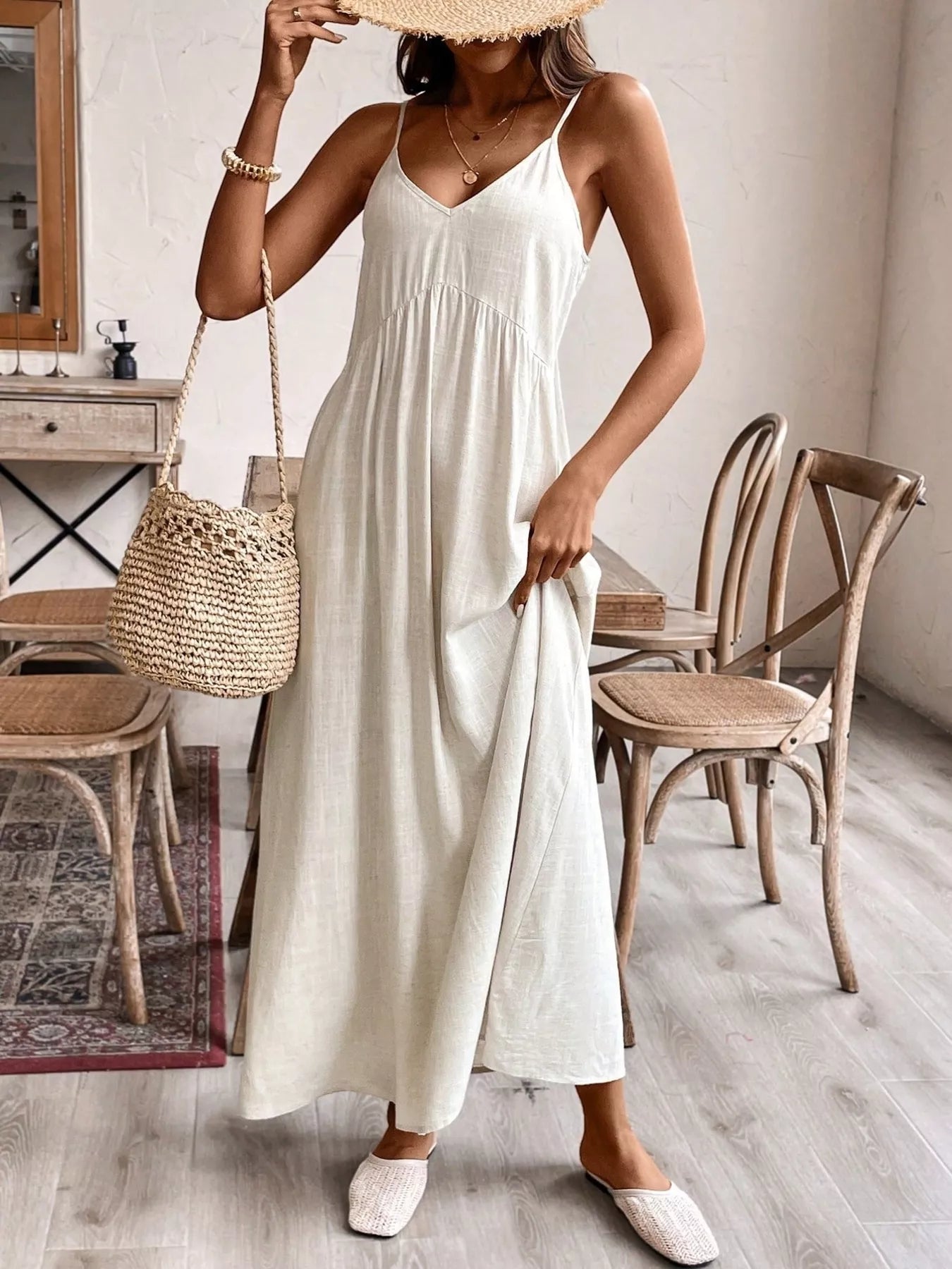 Woman in a white dress holding a straw bag in a room with wooden furniture.