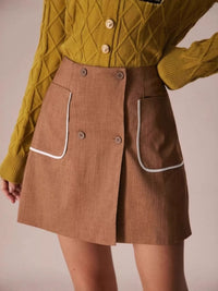 Person wearing a mustard yellow knitted cardigan and brown skirt with white trim on a plain background