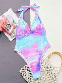 Multicolored tie-dye swimsuit on a gray surface with a pink phone and beige bag.