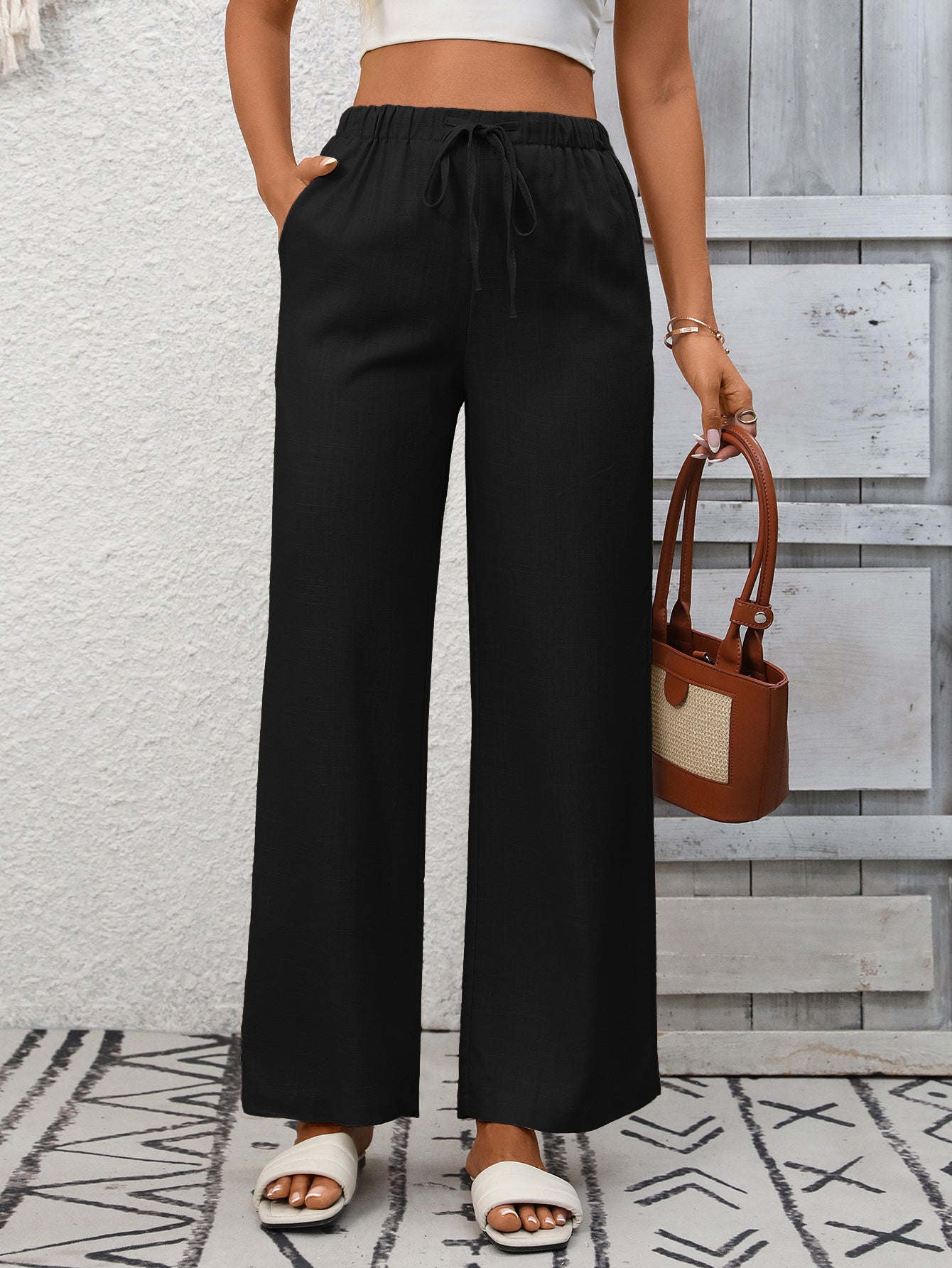 Person wearing black wide-leg pants holding a brown handbag against a light gray wall.