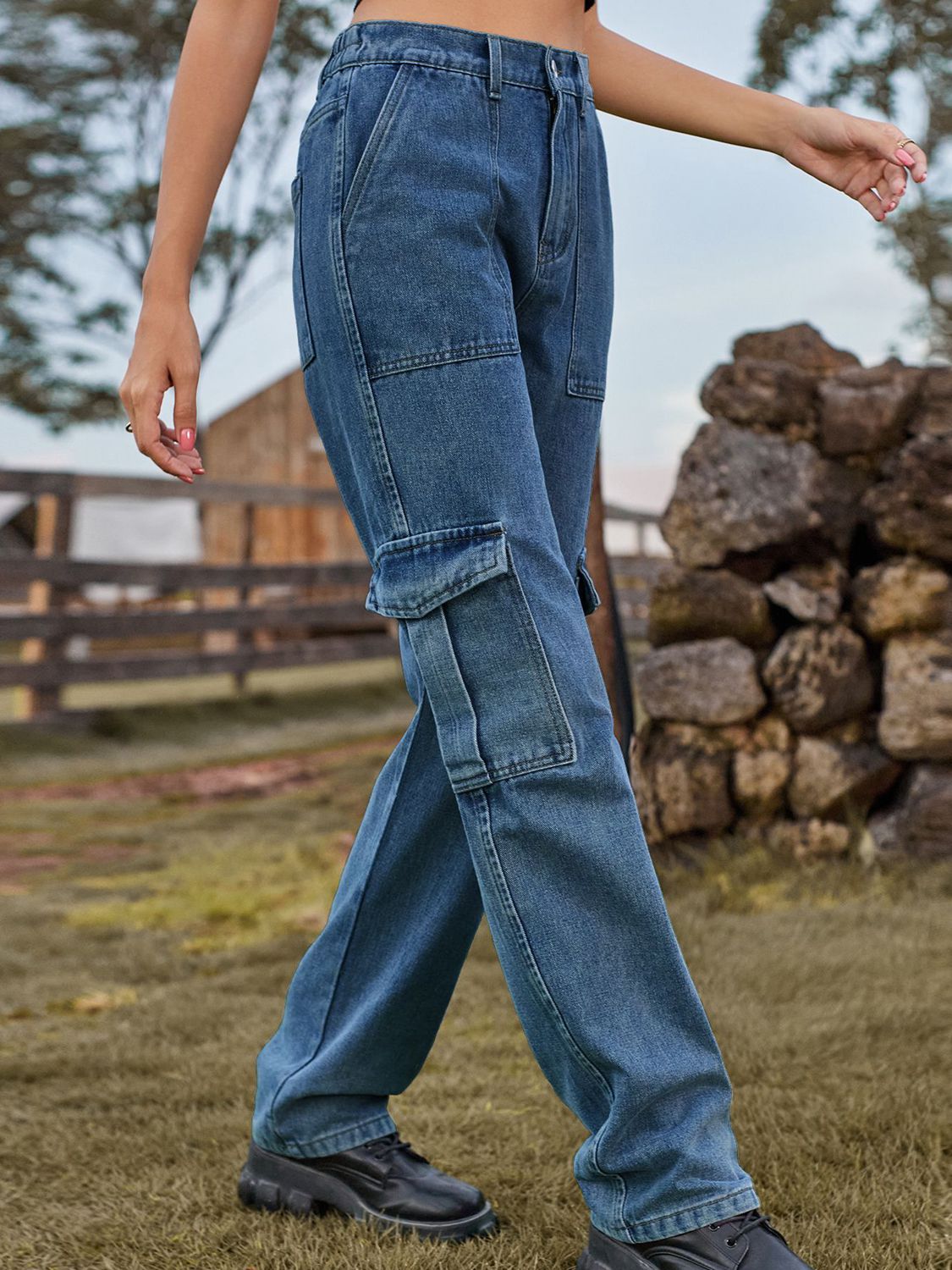 Person wearing blue cargo pants standing outdoors with a wooden fence and stacked logs in the background.