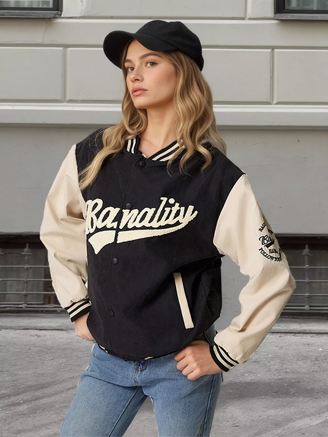 A woman wearing a varsity-style jacket with cream sleeves and collar, black body, and cream script lettering that reads 'Banalité'. She is also wearing a black cap and blue jeans, standing outdoors against a gray building wall.