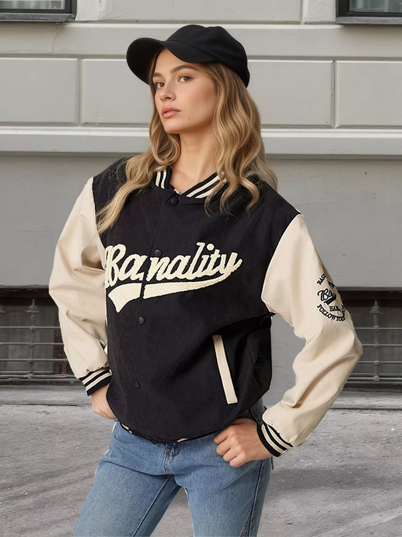 A woman wearing a varsity-style jacket with cream sleeves and collar, black body, and cream script lettering that reads 'Banalité'. She is also wearing a black cap and blue jeans, standing outdoors against a gray building wall.