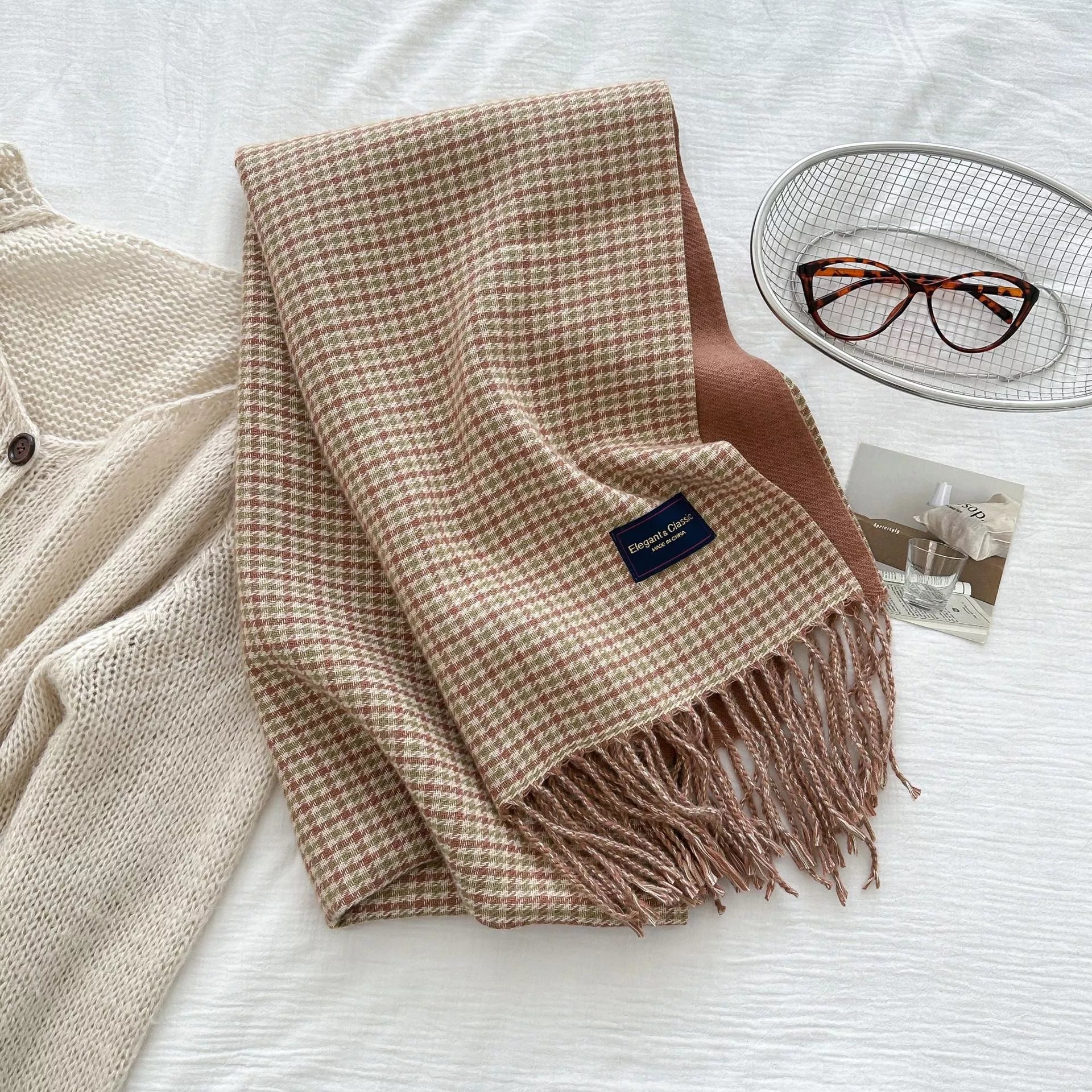Plaid blanket with tassels on a white surface, next to a glass of water and a pair of glasses.