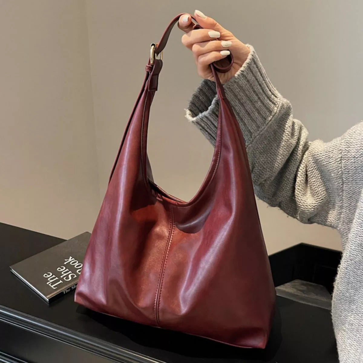 A person holds a slouchy, slouchy leather shoulder bag in a deep burgundy red. The person is wearing a gray knit sweater, and the bag is placed on a black surface next to a book.