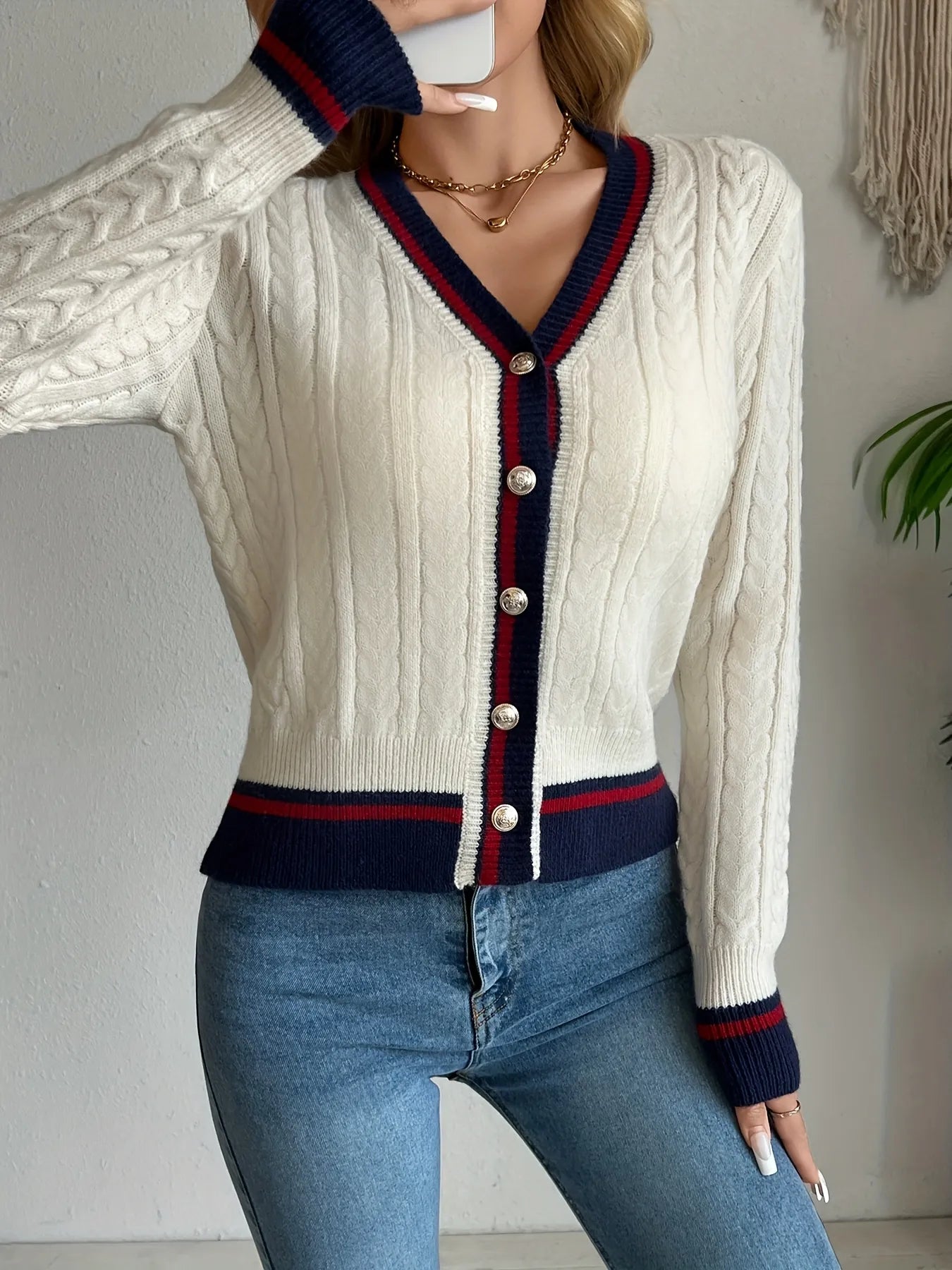 White cable knit cardigan with blue and red trim worn by a person, against a neutral background.