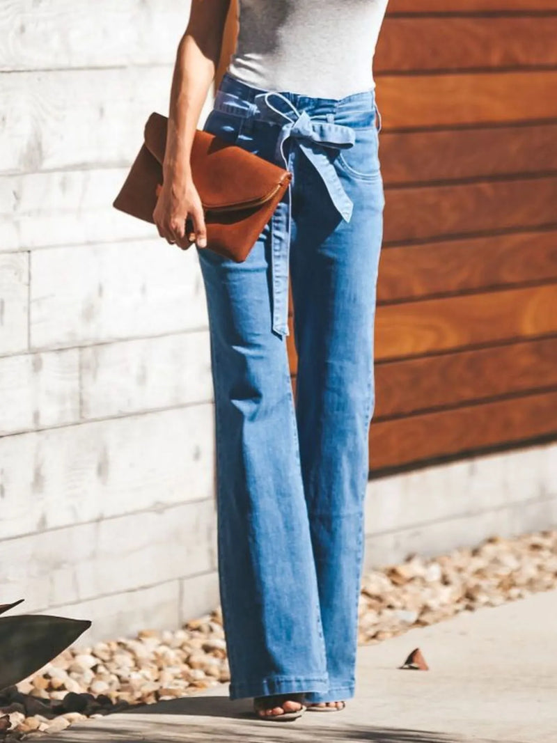 Person wearing blue jeans and a white top, holding a brown clutch against a wooden wall.