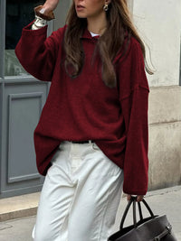 Woman wearing a burgundy sweater and white pants, holding a black handbag, against a building background.