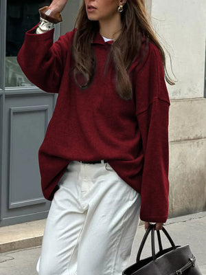 Woman wearing a burgundy sweater and white pants, holding a black handbag, against a building background.