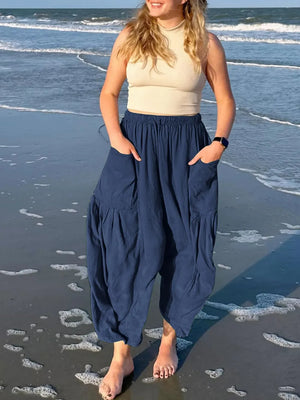 Woman wearing a beige top and blue harem pants standing on a beach.