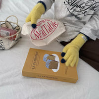 Person wearing yellow gloves holding a 'Meal Table' box on a bed with a 'La Pâtisserie' box.